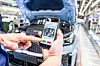Eine Person hält ein Smartphone vor einem Fahrzeug mit geöffneter Motorhaube in einer modernen Autofabrik. Auf dem Smartphone ist eine digitale Qualitätskontrolle-App geöffnet, die Bauteile des Motorraums zeigt. Die Person tippt auf einen grünen Button zur Freigabe. Im Hintergrund ist die Produktionslinie sichtbar.