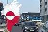 Eine Straßenansicht in einer grönländischen Stadt mit modernen Gebäuden und mehreren fahrenden Autos. Links im Bild ist die Silhouette von Grönland mit der rot-weißen Nationalflagge überlagert. Im Hintergrund stehen blau gestrichene Häuser mit schwarzen Dächern.