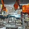 Mehrere orangefarbene Industrieroboter bearbeiten eine schwarze Cockpiteinheit in einer Fertigungszelle. Das Bauteil ist auf einem Gestell eingespannt, während Kamerasysteme und Werkzeuge über dem Werkstück positioniert sind. Im Hintergrund sind verglaste Sicherheitswände und weitere Robotersysteme sichtbar.
