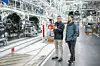 Michael Frieß, Standortverantwortlicher und Leiter der Produktion im Mercedes-Benz Werk Bremen, führt Redakteur Timo Gilgen („Automobil Produktion“) durch die Produktionshalle. Beide tragen wetterfeste Jacken; Frieß erklärt gestikulierend einen Abschnitt der Fertigungslinie, während Gilgen aufmerksam zuhört. Im Hintergrund sind teilweise montierte Fahrzeuge an der Montagelinie sowie moderne Produktionsanlagen und Fördertechnik zu sehen.