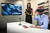 Ein Mann sitzt am Tisch und trägt eine VR-brille. Darüber kann er virtuell sein Wunschfahrzeug ansehen. Daneben steht eine Frau und berät ihn. Über einen Bildschirm kann sie mitverfolgen welche Bilder der Kunde gerade sieht.