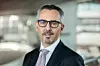 Porträt von Joachim Post, BMW-Vorstand für Einkauf und Lieferantennetzwerk, mit grauem Anzug, weißem Hemd und Krawatte. Er trägt eine Brille, blickt direkt in die Kamera, Hintergrund: moderne Architektur der BMW Welt in München.