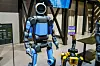 Alt-Tag:
Ein humanoider Roboter von Boston Dynamics steht auf einer Messe- oder Ausstellungsfläche in einer modernen Industriepräsentation. Die Maschine ist in Blau und Dunkelgrau gestaltet, besitzt einen runden, schwarzen Sensorkopf mit kamerähnlicher Front, einen kompakten Oberkörper mit sichtbar segmentierten Gelenken sowie zwei Arme mit mehrgliedrigen Greifhänden und zwei kräftige, menschlich anmutende Beine. Auf der Brust ist der Schriftzug „Boston Dynamics“ zu erkennen. Rechts neben dem Roboter steht der gelbe Roboter-Hund Spot desselben Herstellers, daneben ein Schild mit der Aufschrift „Best of CES Winner“, was auf die Auszeichnung des gezeigten Robotik-Konzepts auf der Consumer Electronics Show hinweist. Im Hintergrund sind Monitore, schwarze Stahlträger, Tische und weitere Präsentationselemente zu sehen, die den Eindruck eines professionellen Technik-Showrooms oder Messestands verstärken. Das Bild visualisiert die im Text beschriebene Entwicklung humanoider Roboter für industrielle Anwendungen bei Hyundai, insbesondere den Einsatz von Boston-Dynamics-Systemen wie Atlas und Spot für anspruchsvolle, monotone, gefährliche und sicherheitsrelevante Aufgaben in der Fertigung und Überwachung.