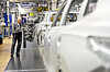 Ein Mitarbeiter arbeitet in der Montagelinie des Volkswagen-Werks Wolfsburg an einer Fahrzeugkarosserie. Im Vordergrund ist unscharf eine weitere Karosserie zu sehen, im Hintergrund Anlagen und Produktionsausrüstung.