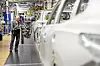 Ein Mitarbeiter arbeitet in der Montagelinie des Volkswagen-Werks Wolfsburg an einer Fahrzeugkarosserie. Im Vordergrund ist unscharf eine weitere Karosserie zu sehen, im Hintergrund Anlagen und Produktionsausrüstung.