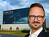 A man in glasses stands in front of a large BMW X5 billboard outside a fenced industrial site.