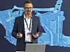 A man in a suit speaks in front of a large blue automotive technical graphic.
