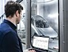 Ein Mann steht vor einer Glasscheibe und blickt auf eine Maschine in einer hellen Produktionshalle.