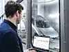Ein Mann steht vor einer Glasscheibe und blickt auf eine Maschine in einer hellen Produktionshalle.