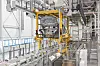Fahrzeugkarosserie in der Montage des BMW Werks Regensburg, aufgehängt in einem gelben Handlingsystem über einer automatisierten Produktionslinie mit Fördertechnik, Schutzgittern und Industrieanlagen für die Fahrzeugfertigung.