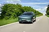 Dunkler SUV auf einer Landstraße mit grünem Straßenrand und Himmel im Hintergrund.