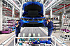 Zwei Beschäftigte stehen vor einem blauen Auto in einer industriellen Montagehalle mit geöffnetem Kofferraum.
