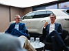 Zwei Männer sitzen vor einem weißen Bentley-SUV in einem modernen Ausstellungsraum und unterhalten sich.