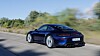 Blauer Porsche-Sportwagen fährt auf einer kurvigen Straße entlang von Bäumen und Gras.