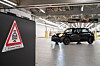 Ein schwarzer Mini fährt in einer hellen Werkhalle an einem Warnschild vorbei.