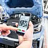 Eine Person hält ein Smartphone vor einem Fahrzeug mit geöffneter Motorhaube in einer modernen Autofabrik. Auf dem Smartphone ist eine digitale Qualitätskontrolle-App geöffnet, die Bauteile des Motorraums zeigt. Die Person tippt auf einen grünen Button zur Freigabe. Im Hintergrund ist die Produktionslinie sichtbar.
