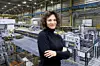 Bilyana Stern, Werkleiterin bei Opel, steht mit verschränkten Armen in einem schwarzen Rollkragenpullover vor dem Hintergrund des Opel-Werks in Kaiserslautern. Im Bildhintergrund sind moderne Presswerk-Anlagen, Metallteile in Gitterboxen und eine helle Industriehalle mit gelben Sicherheitsmarkierungen zu sehen.