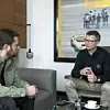 Timo Gilgen, Redakteur der „Automobil Produktion“, im Interview mit Michael Friess, Standortverantwortlicher und Leiter der Produktion des Mercedes-Benz Werks Bremen. Gilgen sitzt links in grüner Cordjacke auf einem grauen Sofa, Friess rechts gegenüber in einem Ledersessel, bekleidet mit beiger Hose und schwarzem Oberteil mit Mercedes-Benz-Logo. Im Hintergrund ein großformatiges Kunstwerk mit Rennwagenszene. Auf dem Glastisch vor ihnen stehen ein Glas Wasser, eine Tasse Kaffee und ein Smartphone.