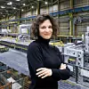 Bilyana Stern, Werkleiterin bei Opel, steht mit verschränkten Armen in einem schwarzen Rollkragenpullover vor dem Hintergrund des Opel-Werks in Kaiserslautern. Im Bildhintergrund sind moderne Presswerk-Anlagen, Metallteile in Gitterboxen und eine helle Industriehalle mit gelben Sicherheitsmarkierungen zu sehen.